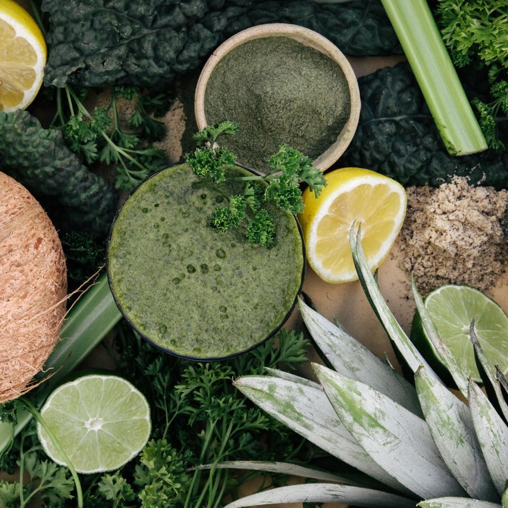 Image of a bird-eye shot of a glass of green smoothie with a parsley garnish beside an open jar with green powder surrounded by cut lemons, cut limes, green vegetables, pineapple, and celery.
