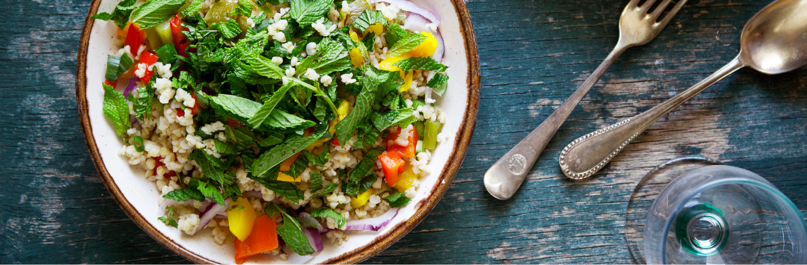 a bowl of salad with quinoa, mint leaves, cilantro, cherry tomatoes, diced red onion and mined garlic beside a glass and a spoon and fork.