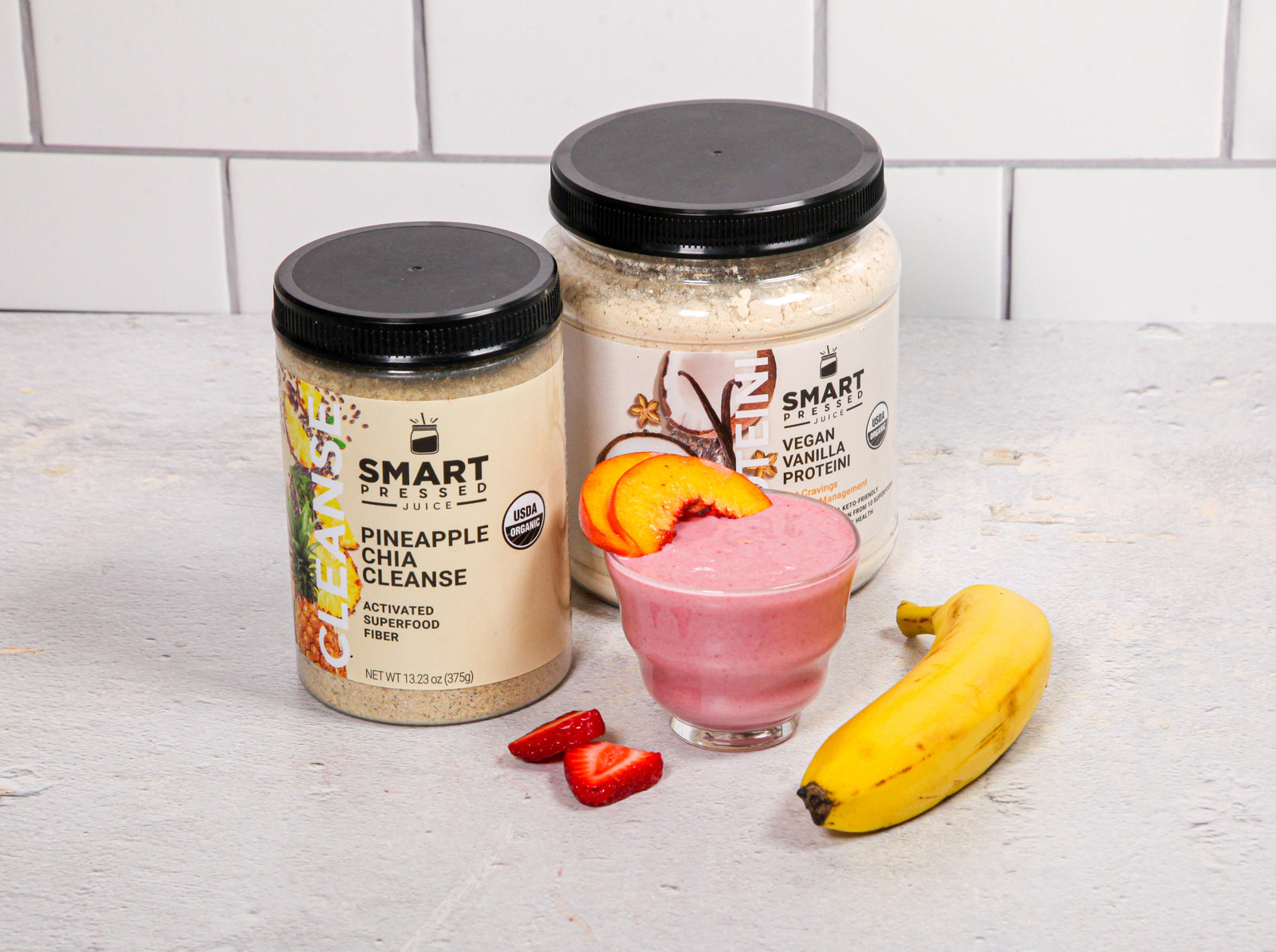 1 jar of Pineapple Chia Cleanse beside 1 jar of Vegan Vanilla Proteini and in front is a glass of Rose's Tropical Proteini topped with slices of apricot beside a banana and slices of strawberry on a marble counter top.
