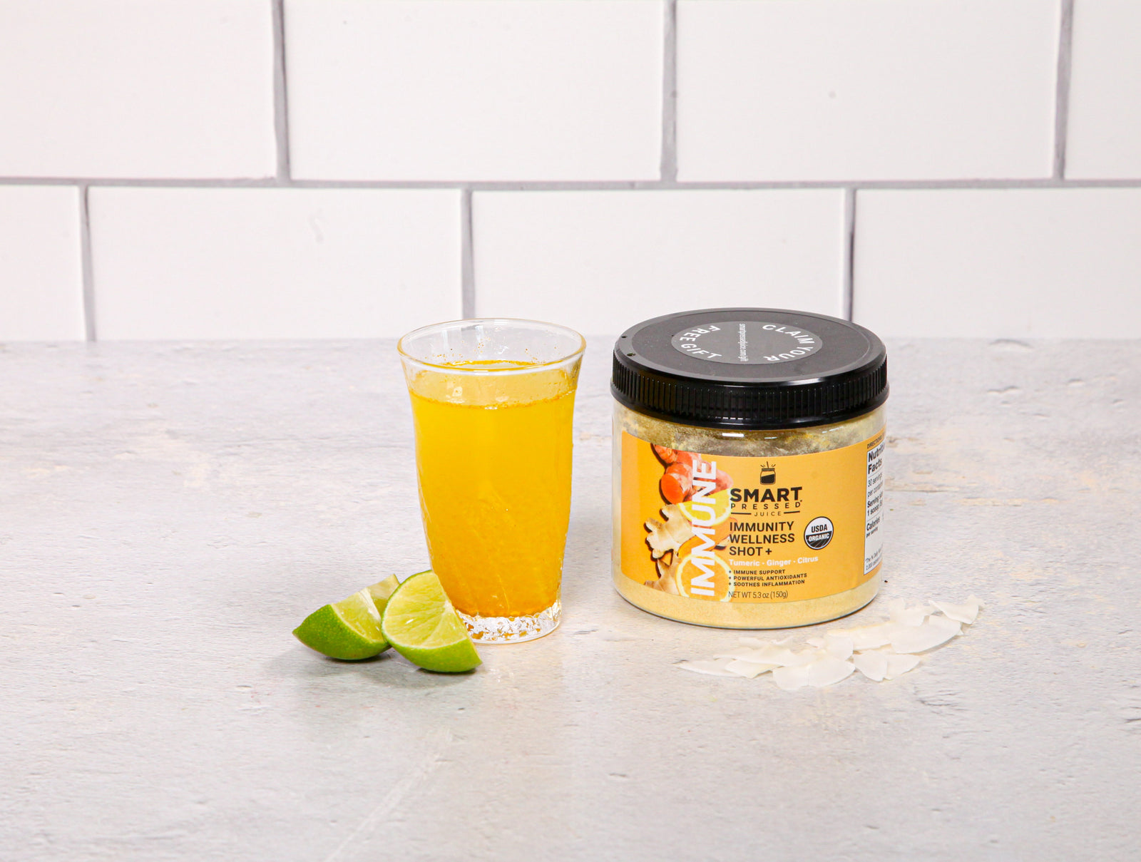 front view of a glass of immunity refresher and a smart pressed immunity wellness shot jar and two wedges of lime on the side on top of a white table