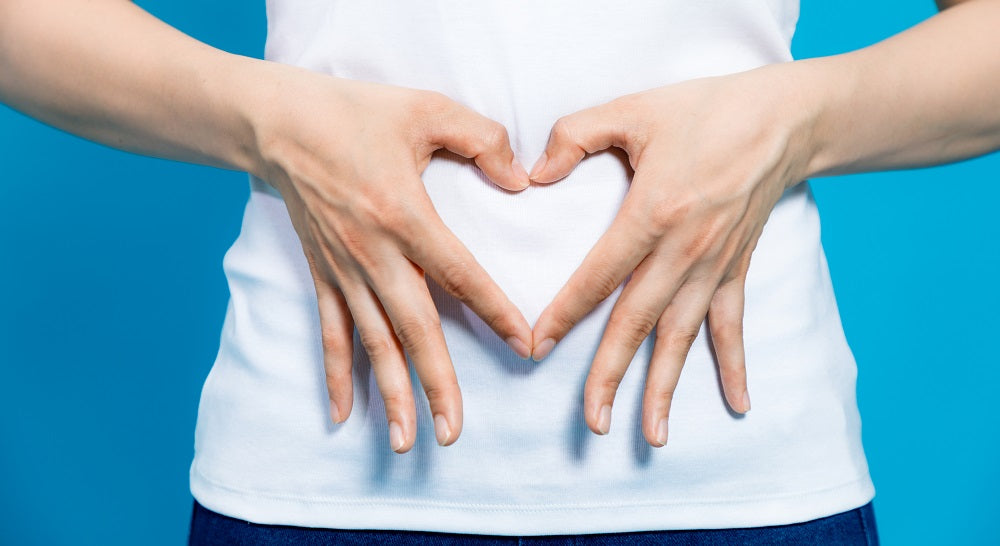 A half-body woman making a heart hand gesture on her lower abdomen standing against a blue wall.