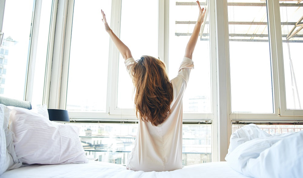 A woman sitting on her bed facing her window while stretching.