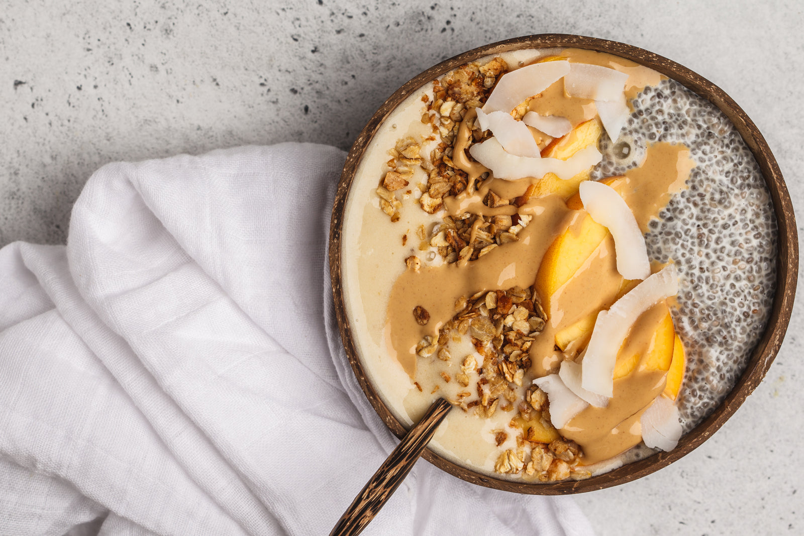 top view of a wooden bowl filled with pineapple chia granola topped with chia seeds, slices of mango, rice flakes, and coconut chips with a wooden spoon and a white cloth on the side on top of a concrete table