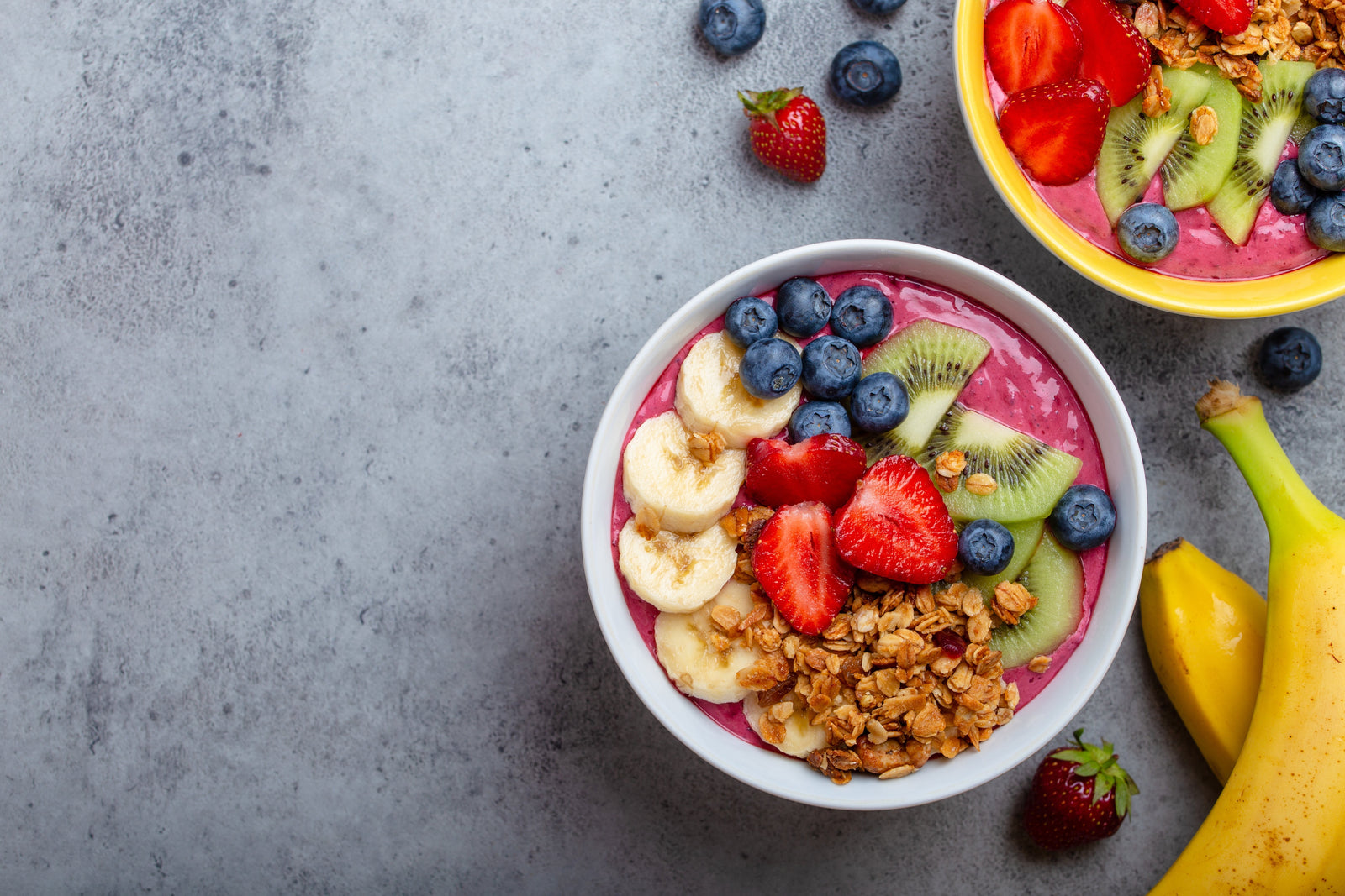 top view shot of 2 bowls of frozen berry smoothie topped with blueberries, sliced strawberries, sliced kiwi, sliced bananas, and granola flakes beside 2 bananas, blueberries, and strawberries.