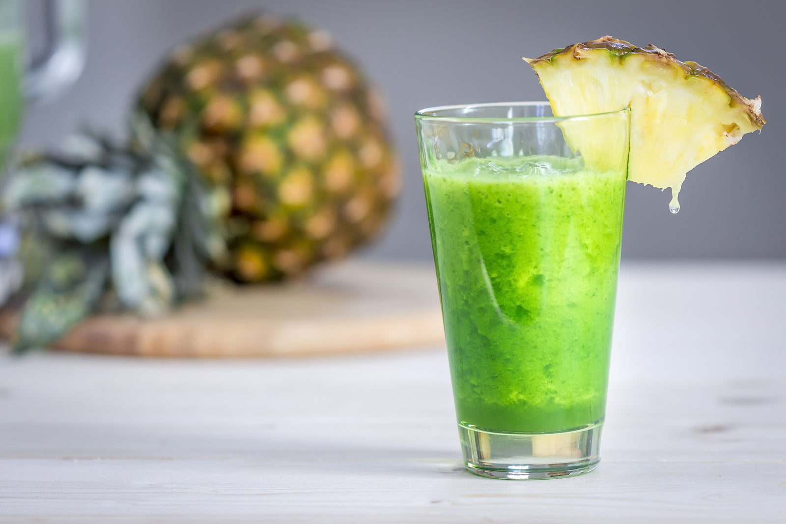 a glass of mixed banana, pineapple, and mango smoothie with a slice of lemon on the of the glass and a half sliced whole pineapple on the side on top of the wooden chopping board