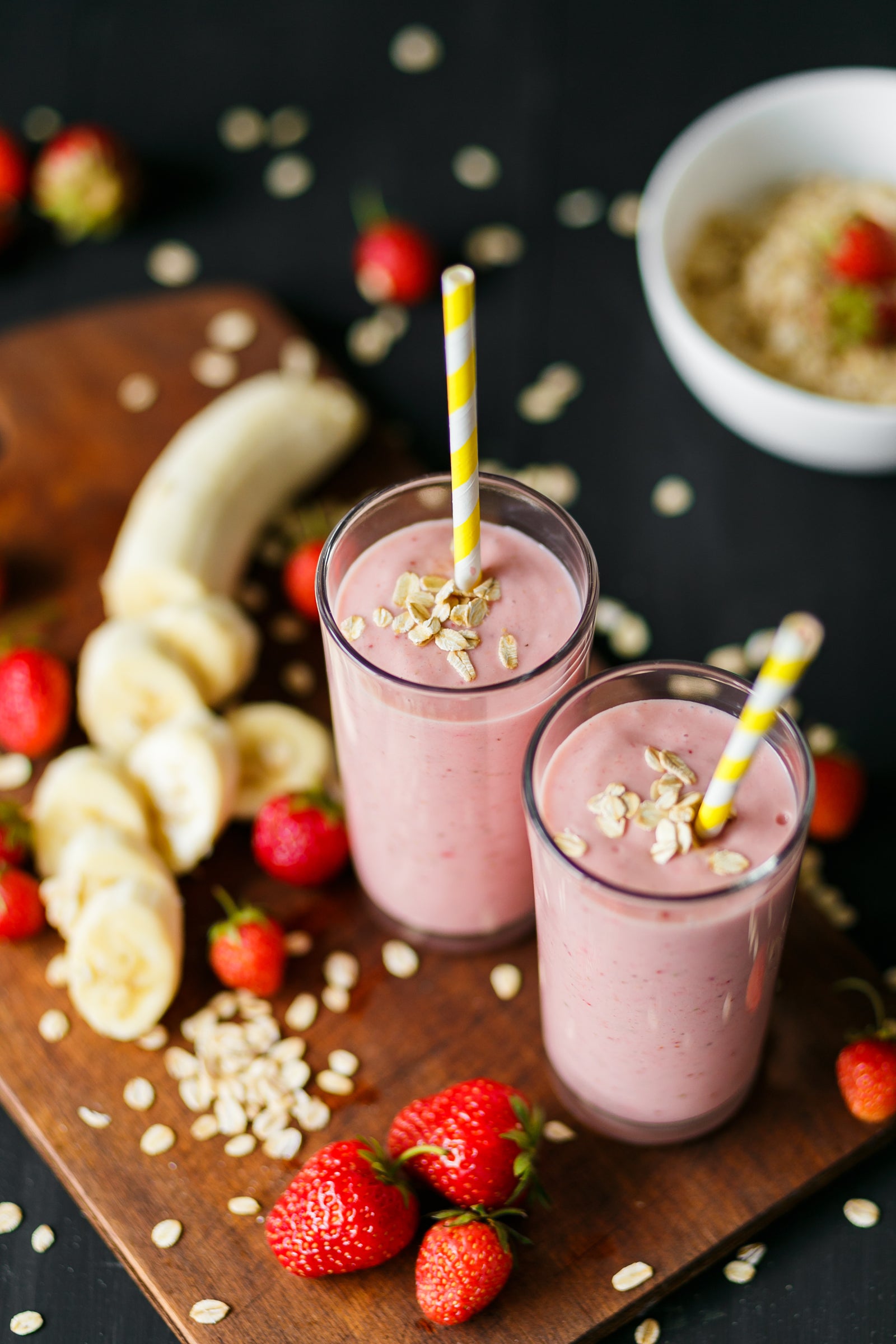 2 glasses of Berry Banana Proteini with straw sprinkled with oats and surrounded with strawberries, oats, and slices of banana on a wooden cutting board.