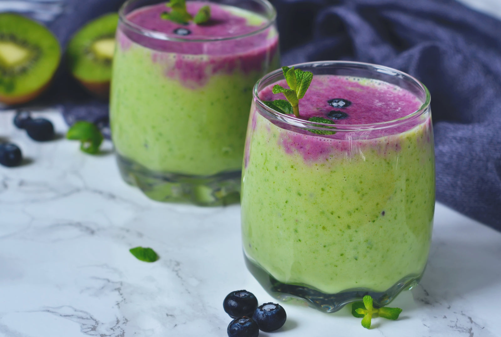 side view of two glasses of pressed greens acai smoothie topped with mint with blue-colored cloth and half-cut kiwi surrounded by blueberries and mint on top of a marble table