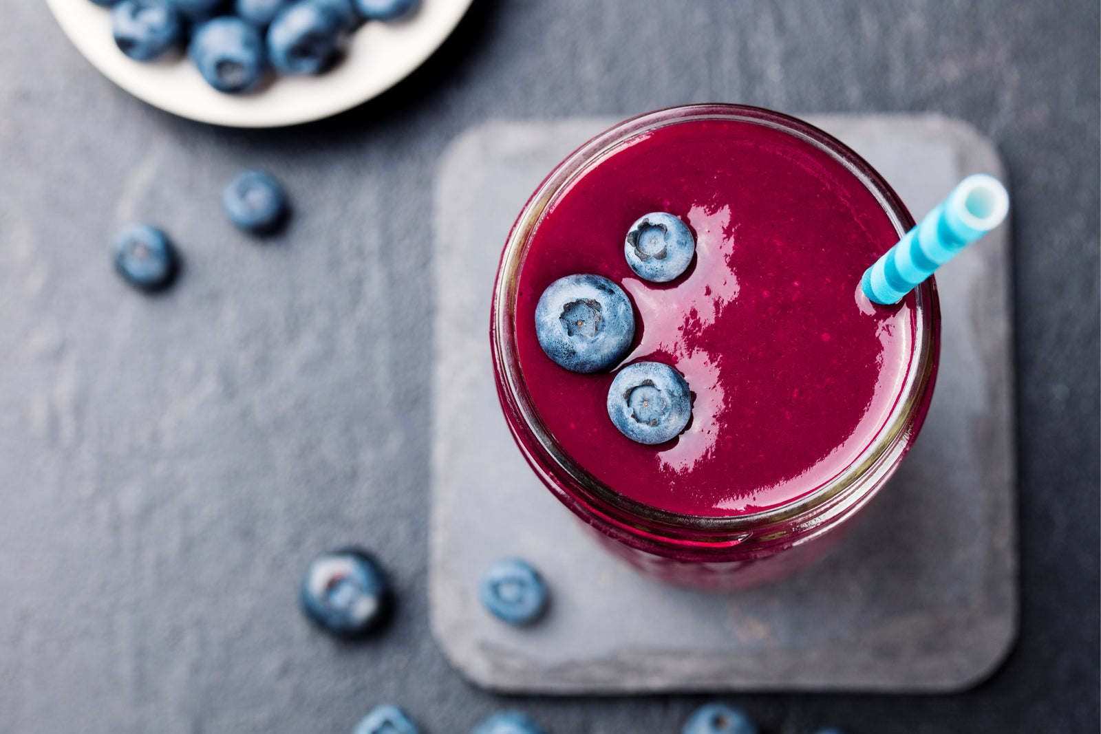 top view shot of a glass of Berry Immunity smoothie topped with 3 blueberries with a straw on a wooden coaster beside a plate of blueberries.