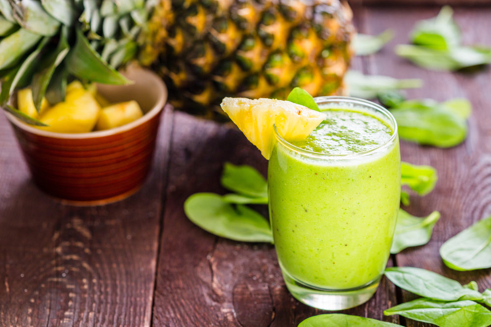 A glass of Pineapple Green Smoothie with a piece of pineapple,, next to a bowl of pineapple and a whole pineapple