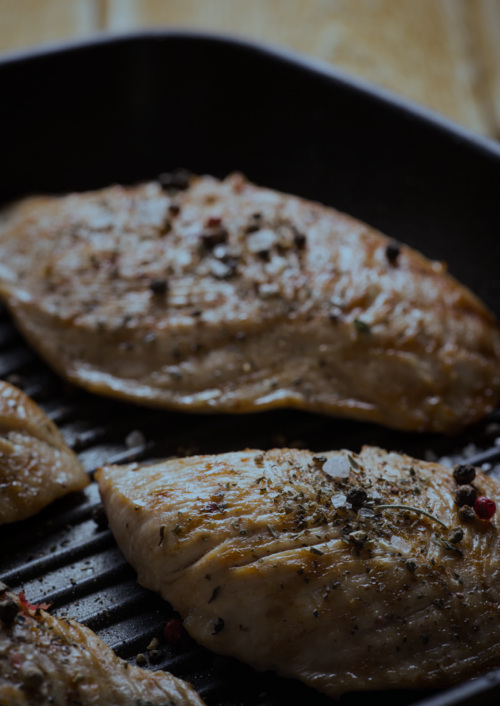 2 chicken breast topped with black pepper corns in a frying pan.