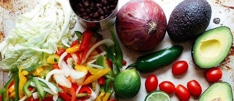 a top view shot of batonnet cut bell peppers and cabbage beside a cup of beans, 1 whole red onion, 1 whole lime and a slice of lime,, 1 green jalapeño, some cheery tomatoes, 1 whole avocado and a half avocado.