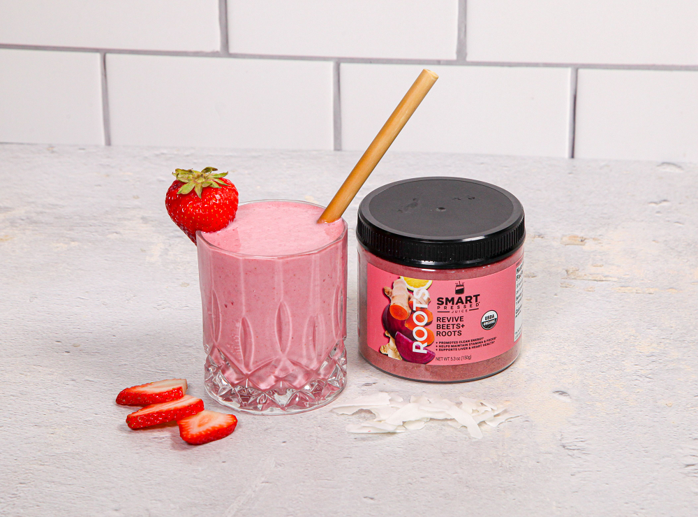 a glass of fruity beets smoothie with the whole strawberry on the side and a plastic brown straw and a smart pressed labeled jar of revive beets roots