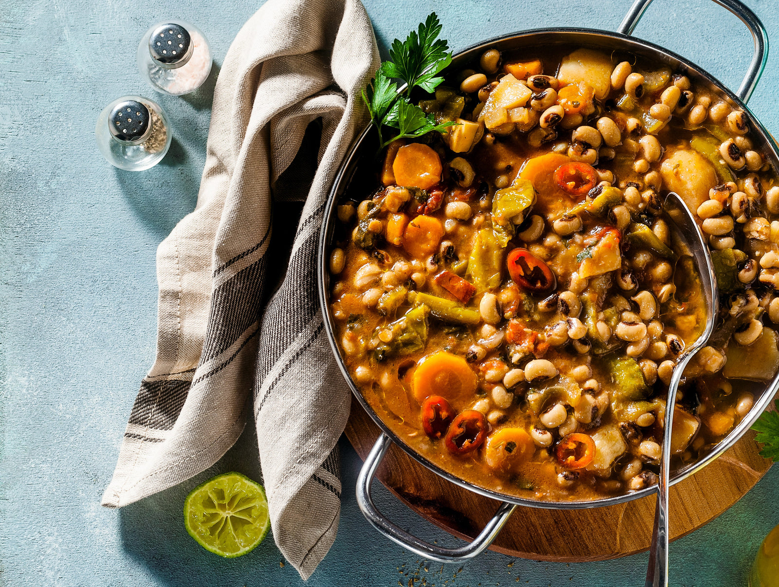 top view of skillet with a stainless spoon on top placed over wooden chopping board filled with black-eyed peas and greens with light brown and dark brown patterned table clothe, half sliced lemon and two condiments bottle on the side of skillet