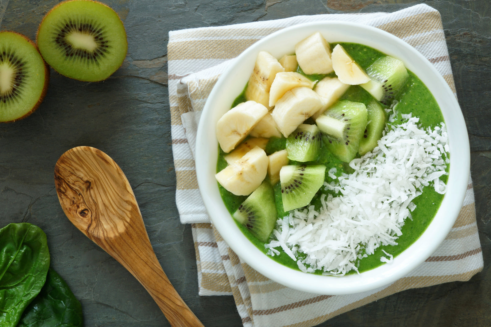 top view of a bowl of coconut water pressed greens smoothie on top of a brown white folded table cloth topped with slices of banana, kiwi, and desiccated coconut surrounded with wooden spoon, arugula, and two slices of kiwi