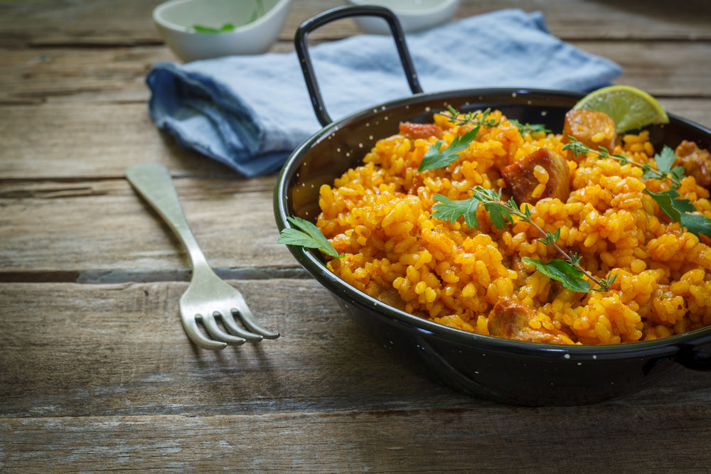 a skillet full of vegetable paella with golden barley topped with five parsley and lemon wedge surrounded by stainless fork, folded blue table clothe and two small white bowl